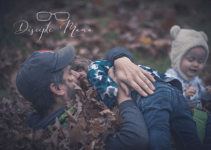 Father lying on the ground in pile of leaves holding son, baby plays in background | Disciple Mama